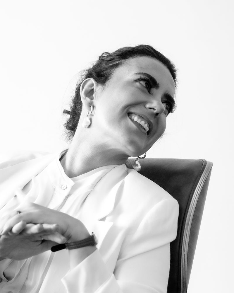 Monica Helena Moreira Pires sorrindo, de blazer branco, sentada numa poltrona